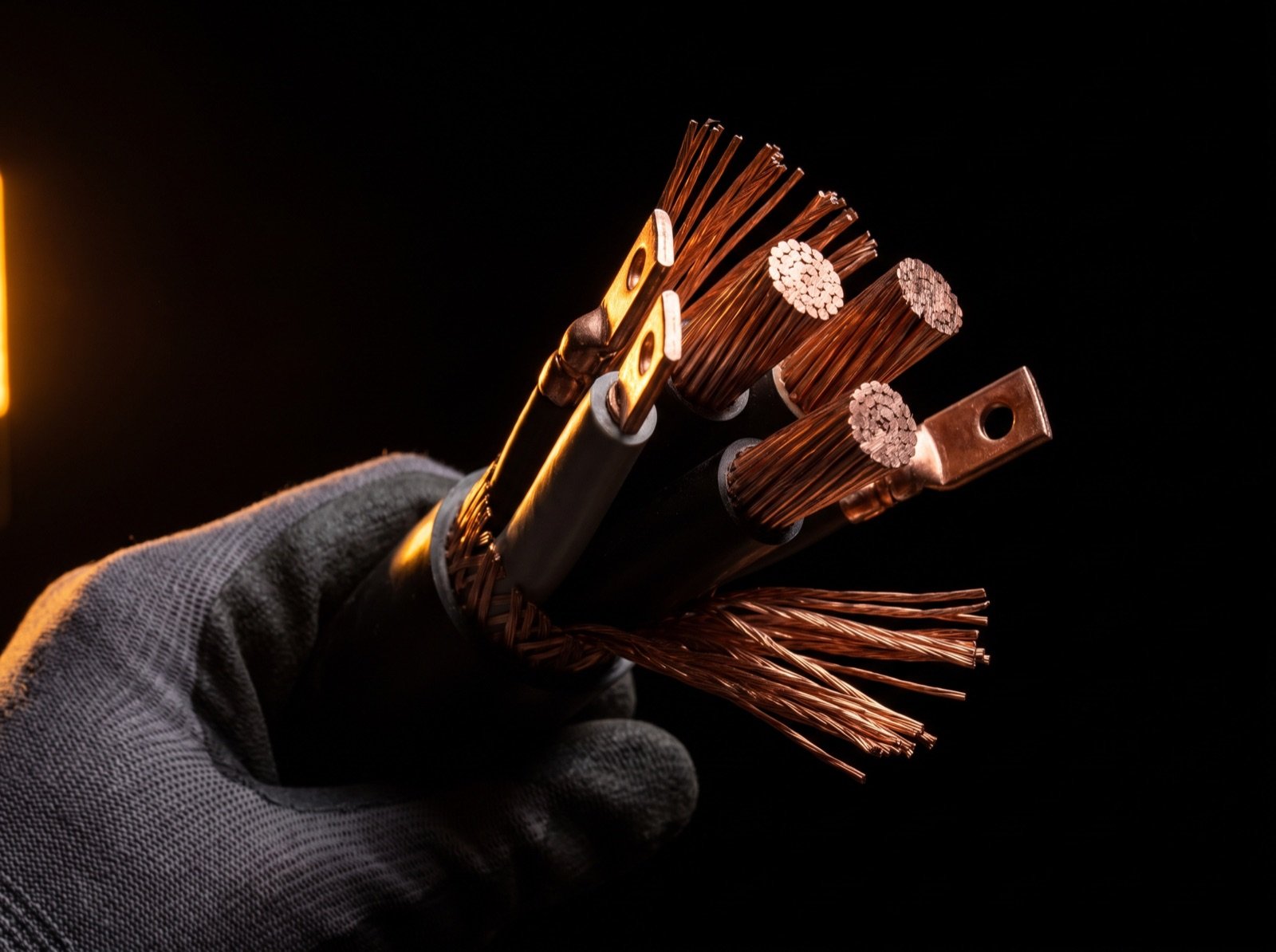 A gloved hand holding a stripped multi-core medium-voltage cable, copper conductors fanned out with crimp lugs fitted, lit by a single amber light against black